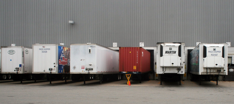 FIGURE 4. Trailer-Type Equipment: Dry Vans, an Ocean Container, and Refrigerated Trailers
This image shows six semi-trailers positioned at a row of warehouse loading bays. Three of the semi-trailers are conventional dry vans, one is a container used in ocean shipping, and two have refrigerator units attached.
